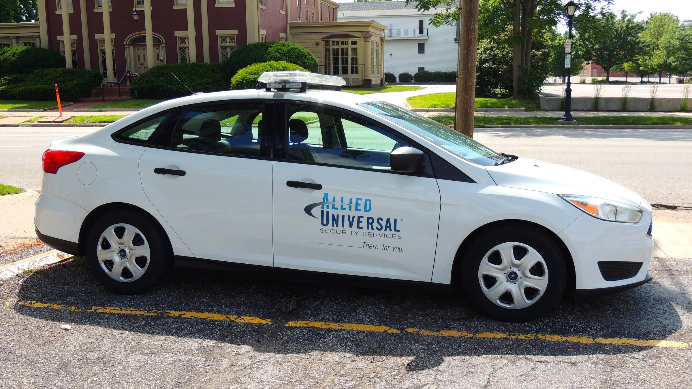 A white Allied Universal Security Services patrol car with an amber light bar on its roof, parked at a curb, with the company's logo and the words "There for you" on the front door