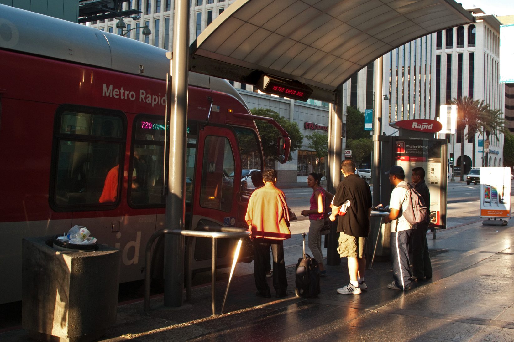 Passengers waiting and boarding a red Metro Rapid 720 bus at the eastbound stop on Wilshire Boulevard at Western Avenue in low evening light