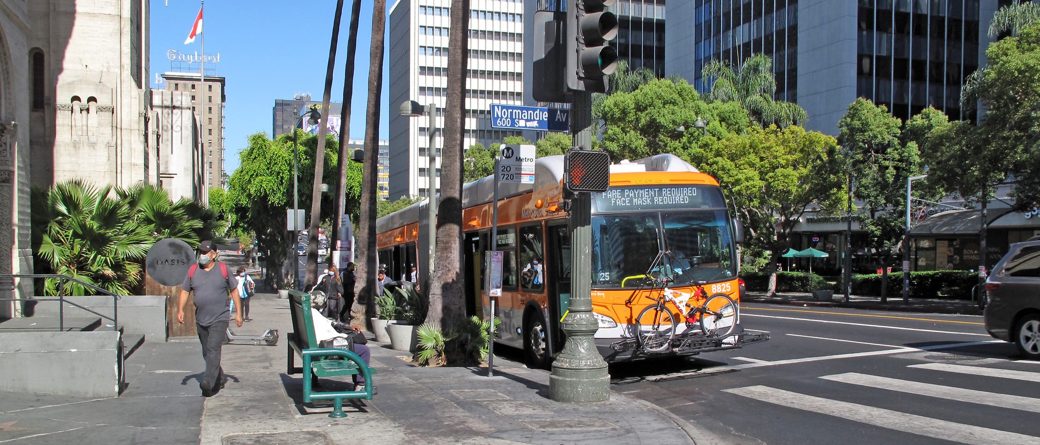 A Metro articulated bus stopped in the curb lane of Wilshire Boulevard at Normandie Avenue, with the stop flag for lines 20 and 720 and the Normandie Avenue street sign