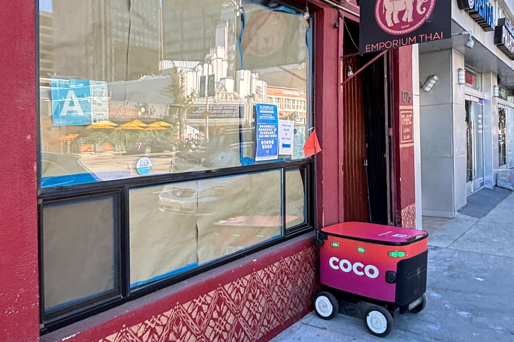 A pink and black Coco delivery robot parked on the sidewalk beside the doorway of Emporium Thai, a restaurant on Westwood Boulevard, under its elephant sign