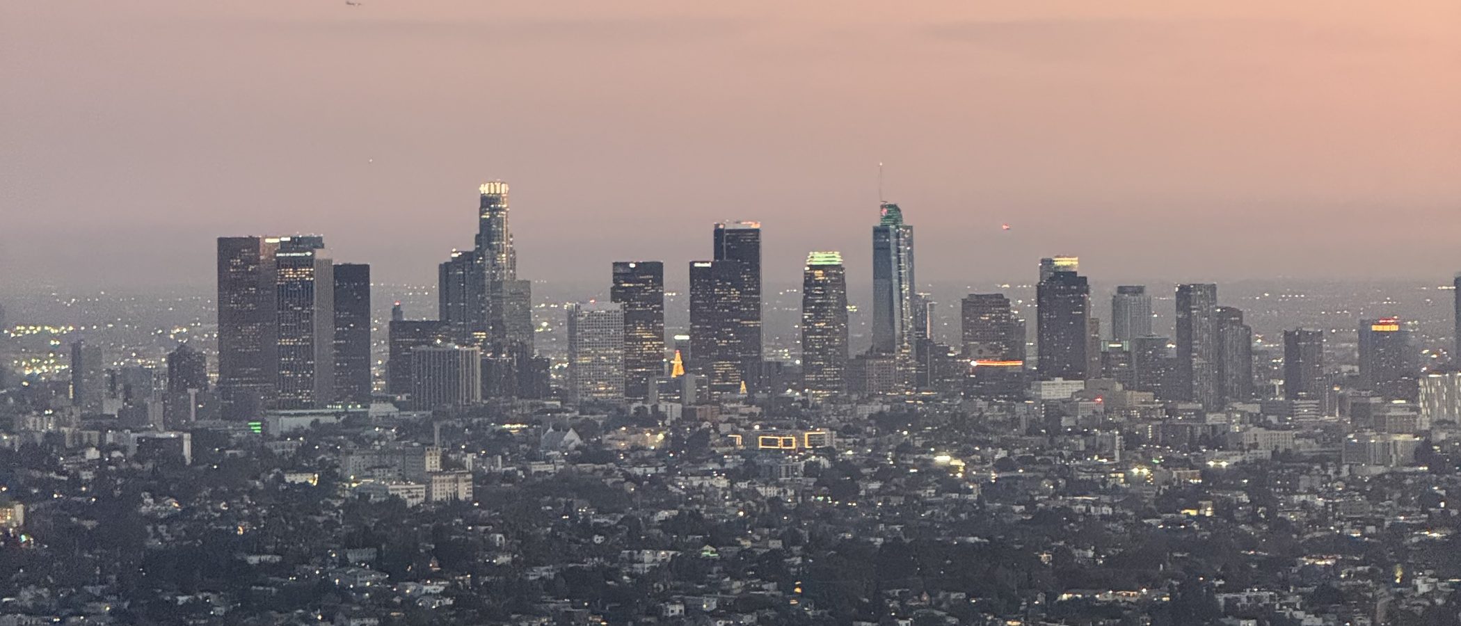 Downtown Los Angeles seen from the hills at dusk, the office towers lit against a pink sky with the rest of the city spread out below them
