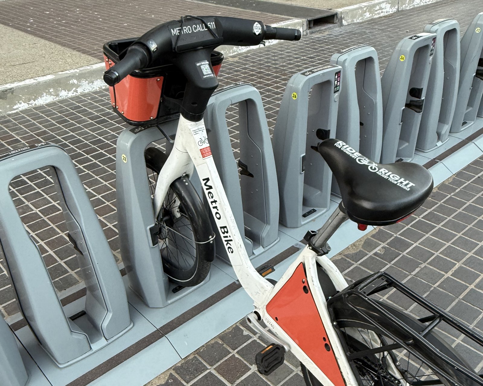 A single Metro Bike Share bicycle docked in a rank of otherwise empty docks on a paved sidewalk in downtown Los Angeles