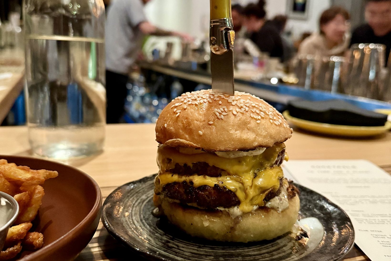 A double cheeseburger on a ceramic plate with a knife stood upright through the bun, a bowl of fries beside it, and the lit counter and diners of a restaurant behind