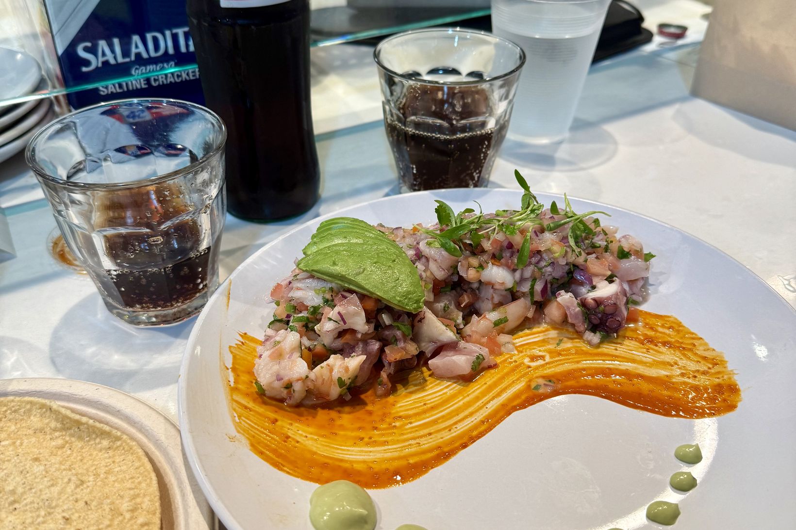A seafood tostada plate with avocado and herbs, a stack of corn tortillas at the edge of the frame, a glass bottle of Coca-Cola and a box of saltines on a white counter
