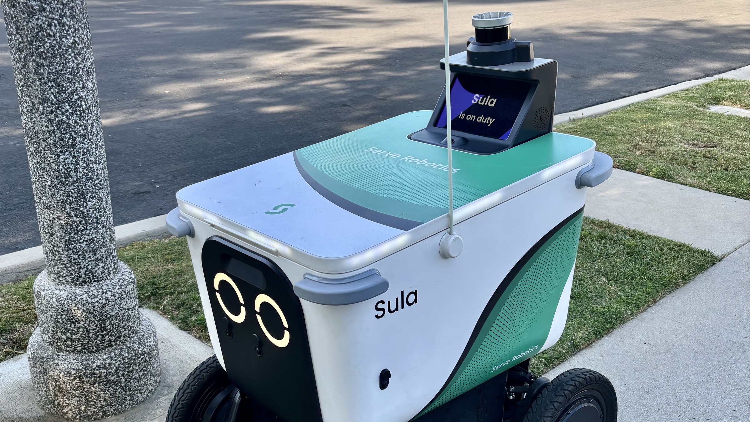 A white and green Serve Robotics delivery robot named Sula on a sidewalk beside a strip of grass, its lit round eyes facing forward and its screen reading "Sula is on duty"