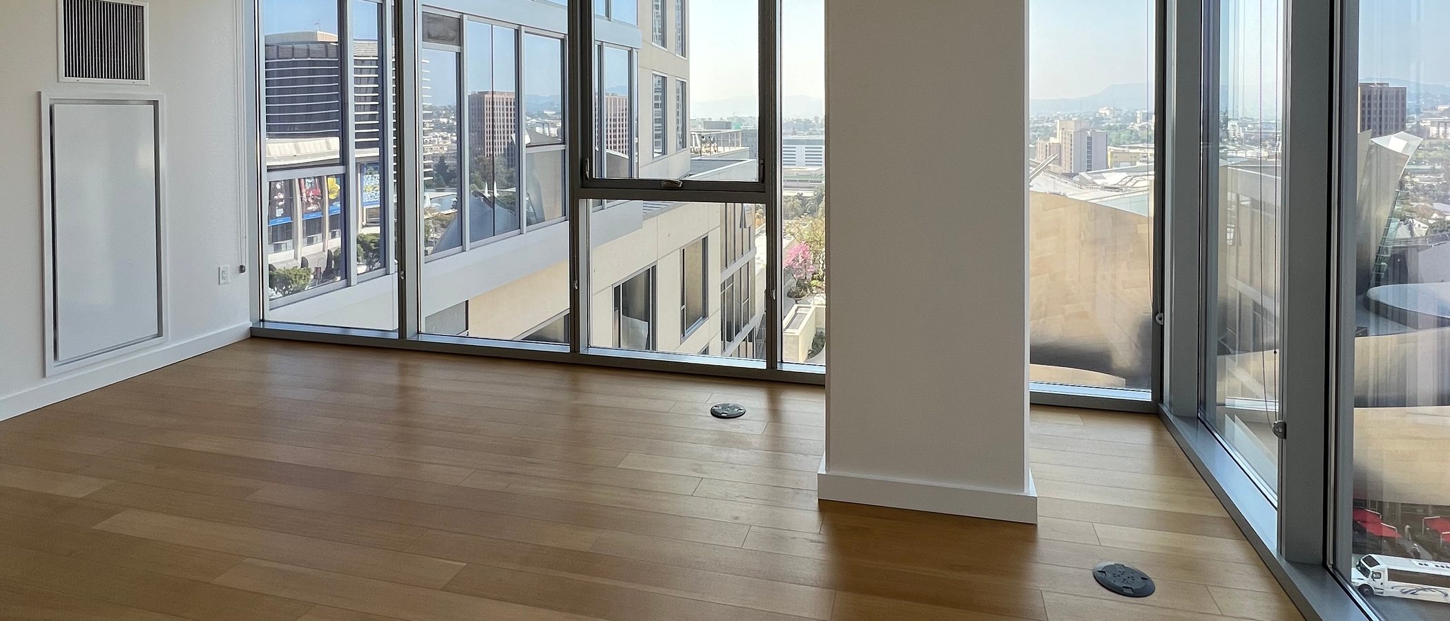 An empty apartment high in a Downtown Los Angeles tower: bare wood floor, white walls and floor-to-ceiling glass on two sides looking out over office buildings, low-rise rooftops and the mountains beyond