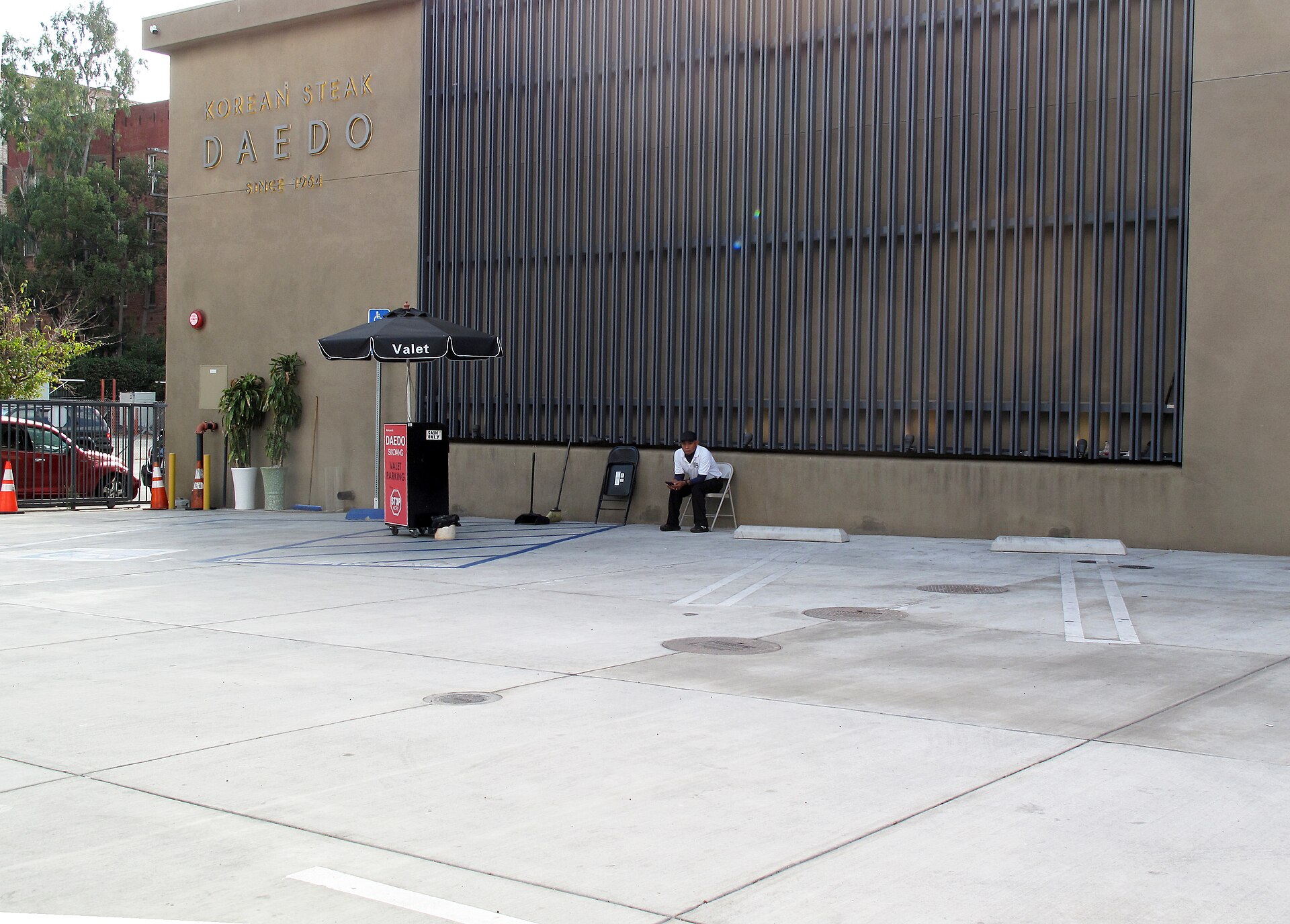 A valet stand under a black umbrella marked Valet in the parking lot of Daedo, a Korean steakhouse in Koreatown, with an attendant seated on a folding chair against the wall and the lot empty
