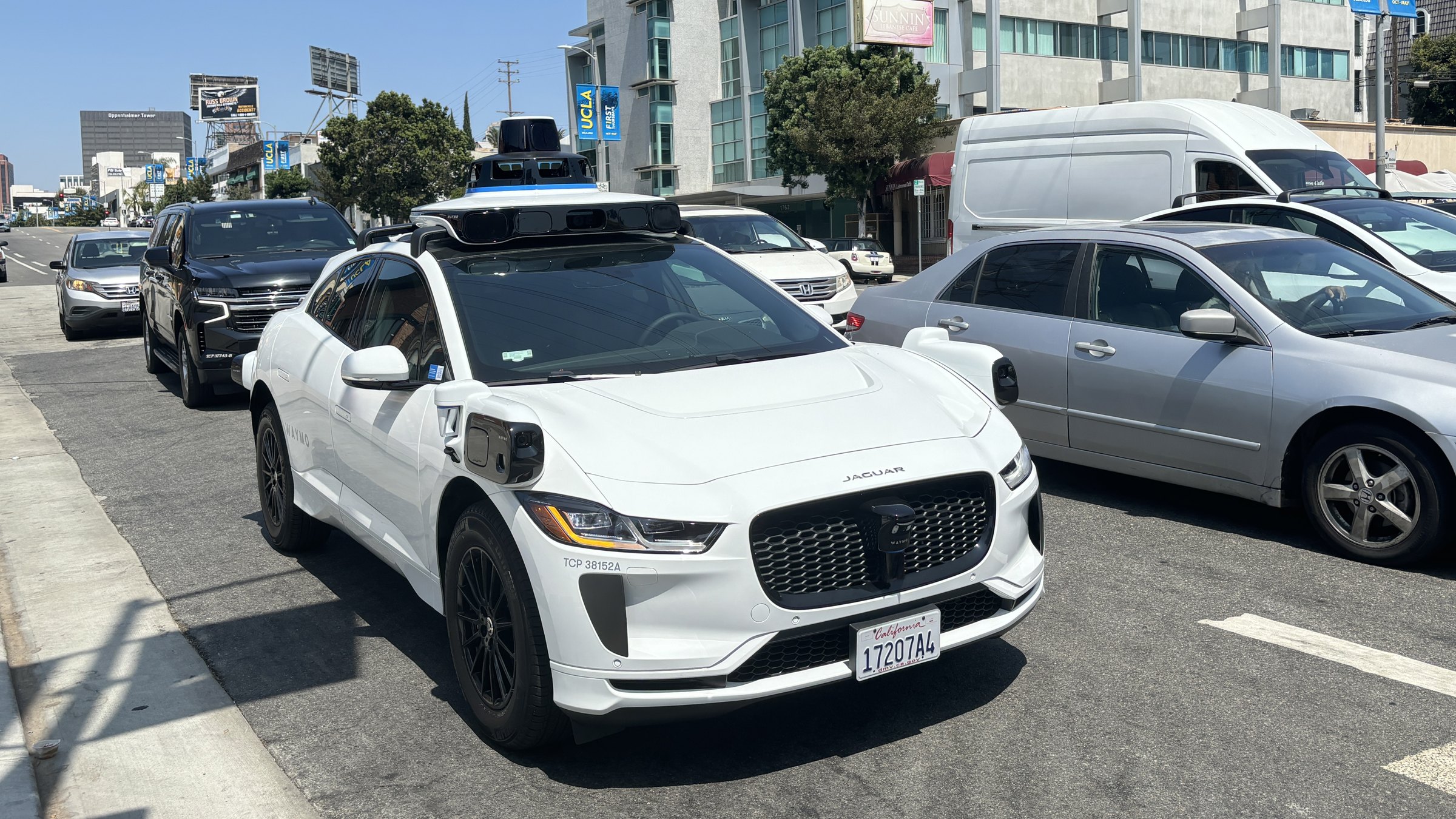 A white Waymo Jaguar with roof sensors stopped in traffic on Westwood Boulevard, with cars beside and behind it and office buildings in the background