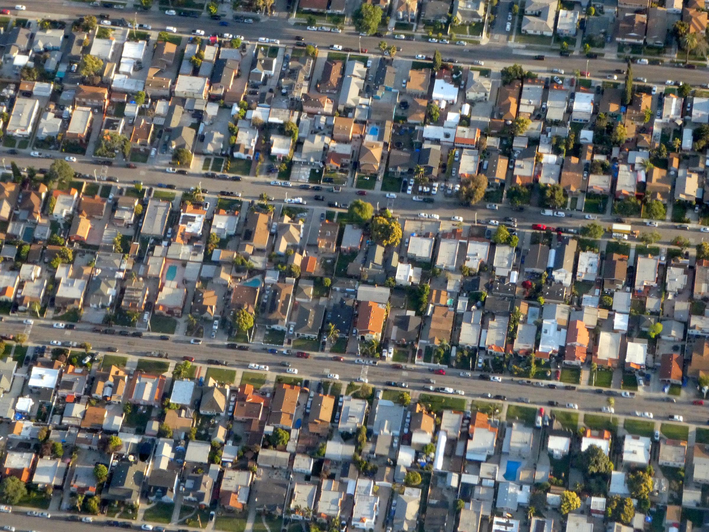 Rows of single-family houses with driveways, backyards and parked cars along three wide streets in Walnut Park, an unincorporated neighborhood of LA County, seen from the air